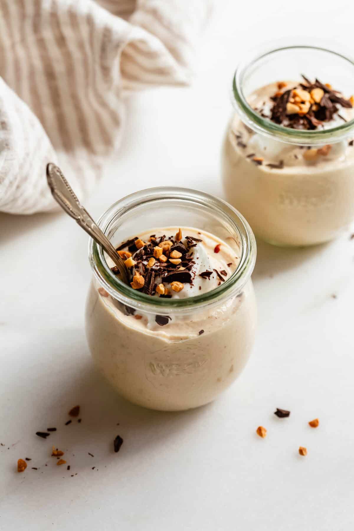 Two jars of Greek yogurt peanut butter mousse with a spoon, garnished with chopped nuts and chocolate.