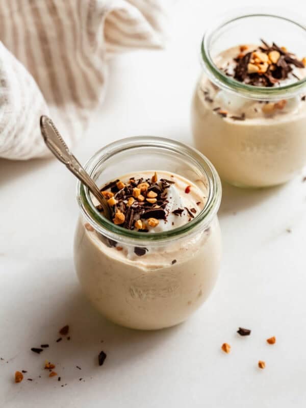 Two jars of Greek yogurt peanut butter mousse with a spoon, garnished with chopped nuts and chocolate.