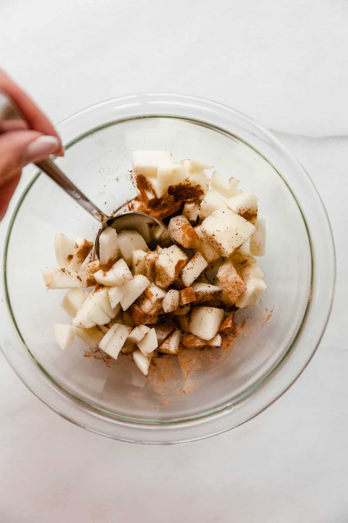 Diced apples being tossed with cinnamon in a glass mixing bowl.