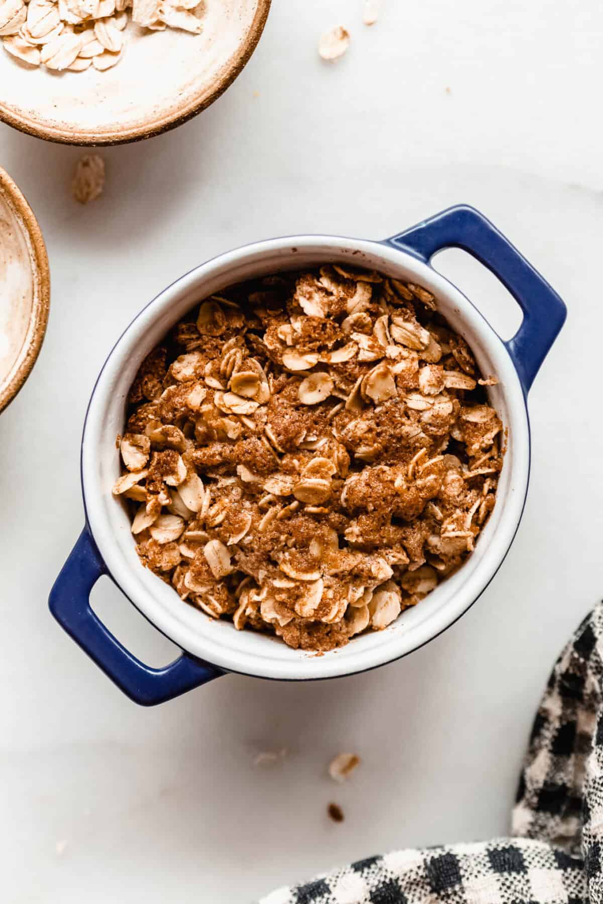 Unbaked apple crisp in a small blue ramekin surrounded by bowls of oats on a marble counter.