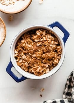Unbaked apple crisp in a small blue ramekin surrounded by bowls of oats on a marble counter.