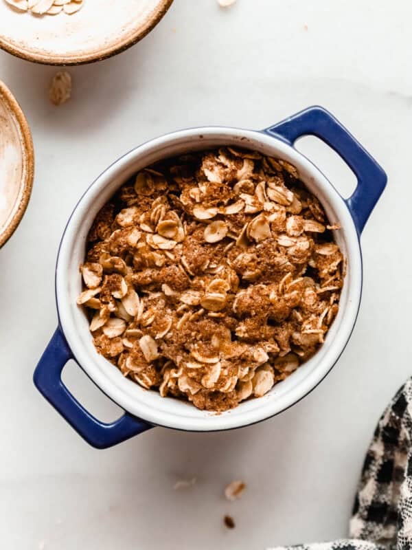 Unbaked apple crisp in a small blue ramekin surrounded by bowls of oats on a marble counter.