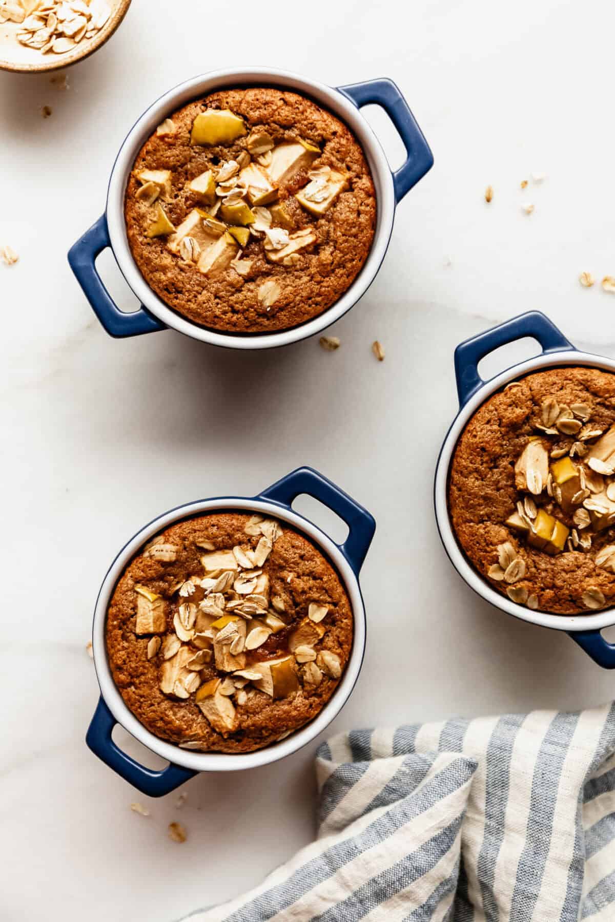 Three apple baked oat ramekins staggered on a marble counter beside a striped linen napkin.