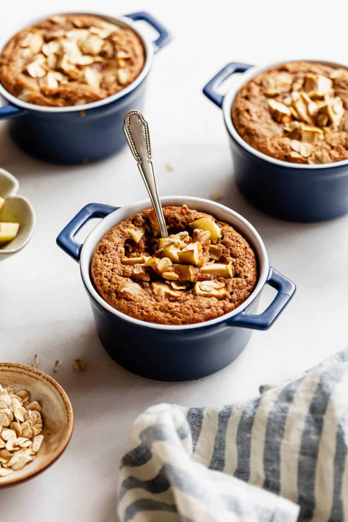 Three individual apple baked oat ramekins arranged together with a bowl of rolled oats nearby.