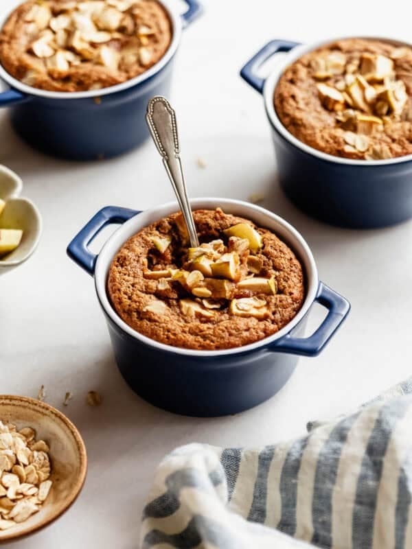 Three individual apple baked oat ramekins arranged together with a bowl of rolled oats nearby.
