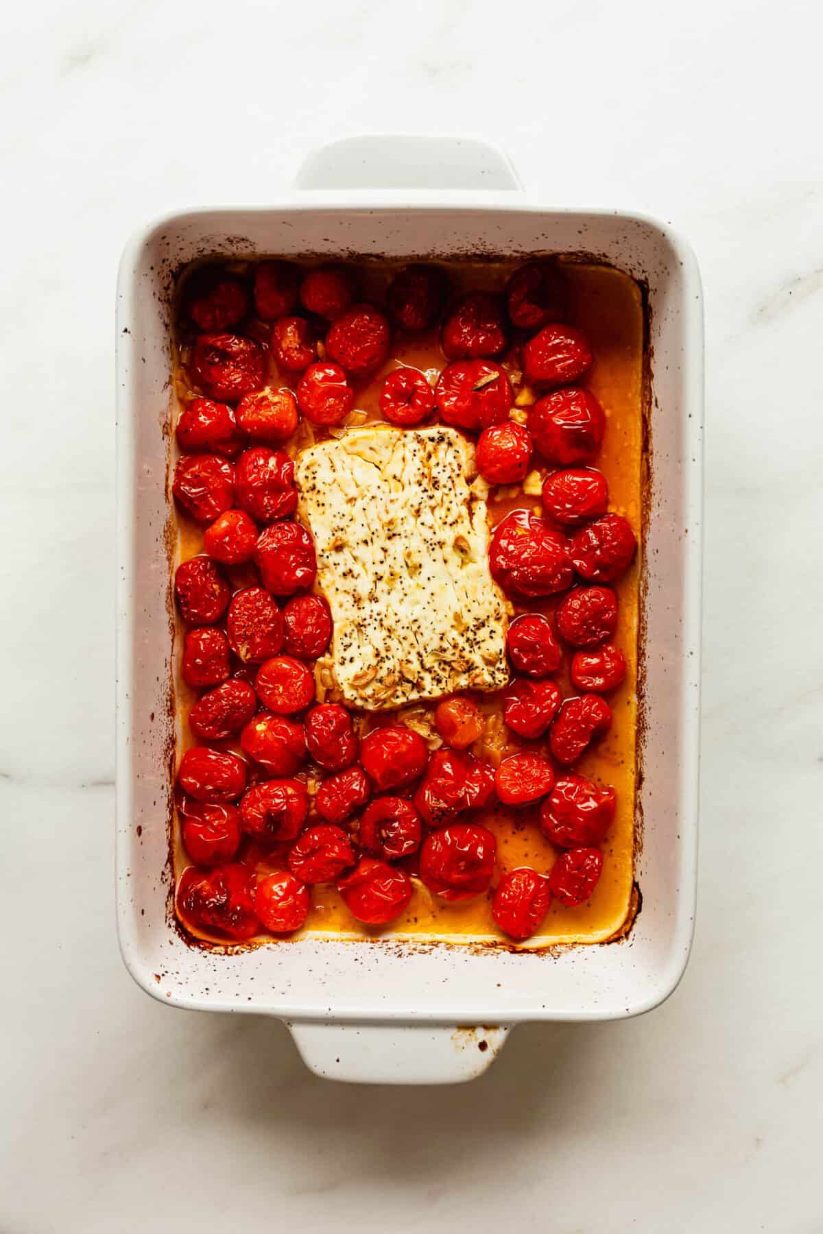 Baking dish of roasted cherry tomatoes surrounding a block of feta cheese.