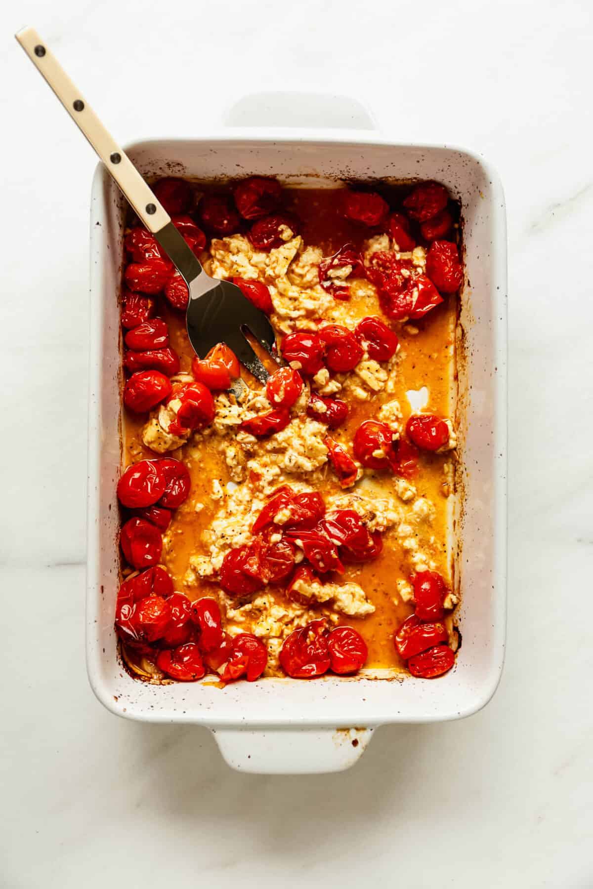 Mashing baked feta cheese with roasted cherry tomatoes using a fork.