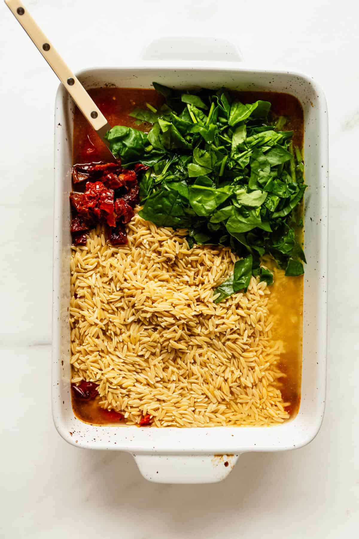 Baking dish of orzo mixed with spinach and sun-dried tomatoes being stirred with a wooden spoon.
