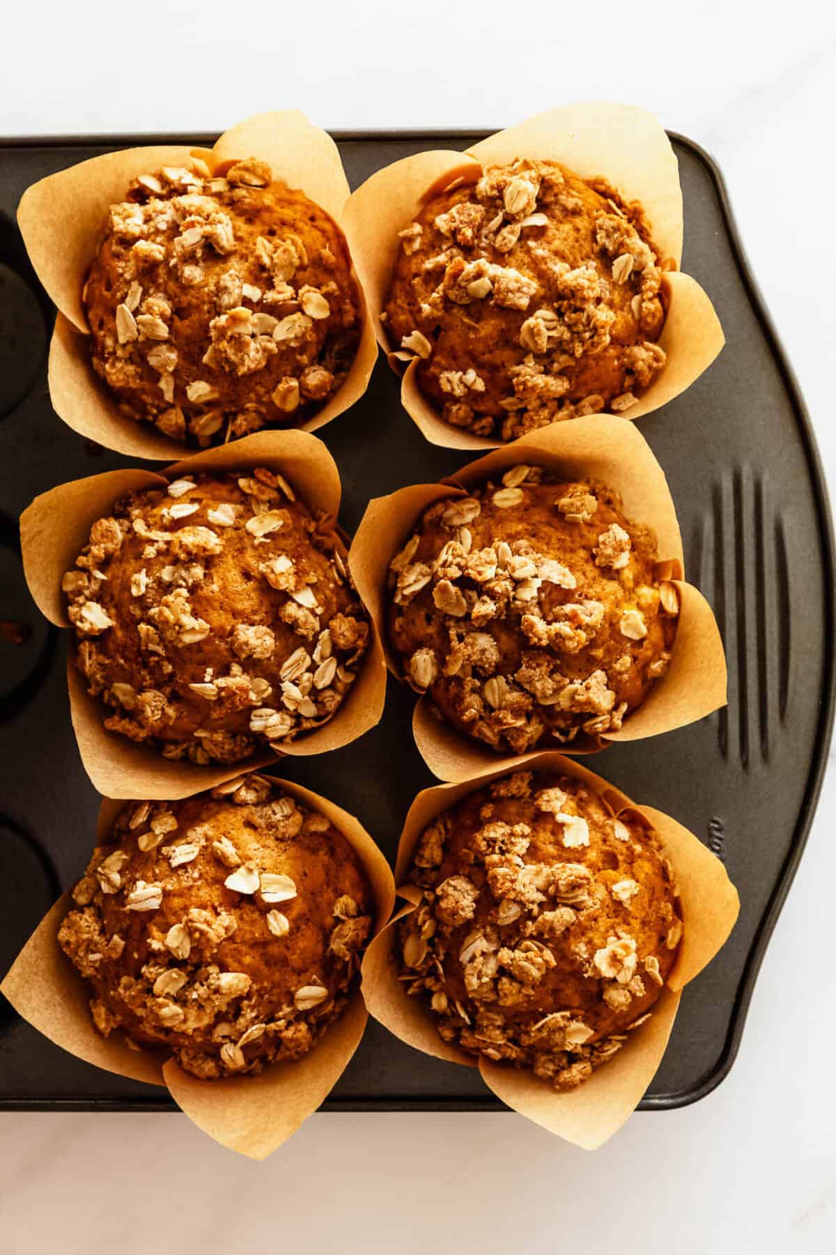 Muffin tin with six banana pumpkin muffins topped with oats, viewed from above.