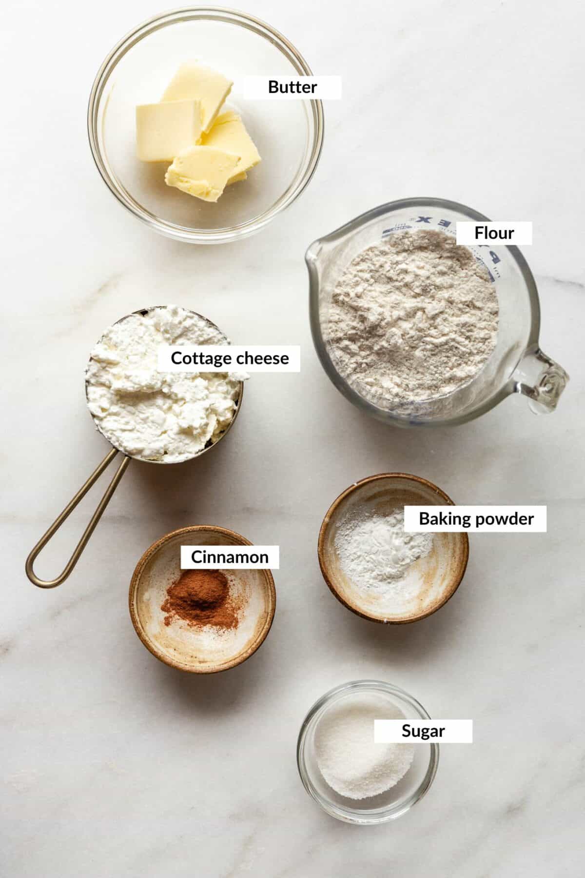 Bowls of butter, flour, cottage cheese, baking powder, cinnamon, and sugar labeled as ingredients for cottage cheese donut holes.