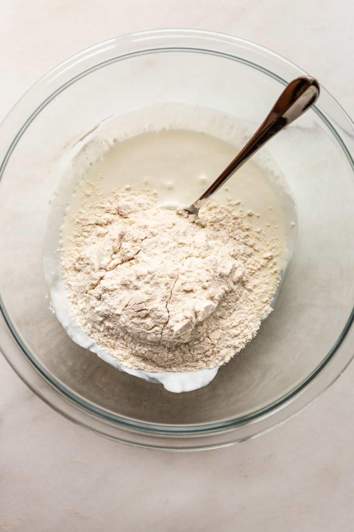 A glass bowl of flour with cottage cheese being stirred together with a spoon to form dough.