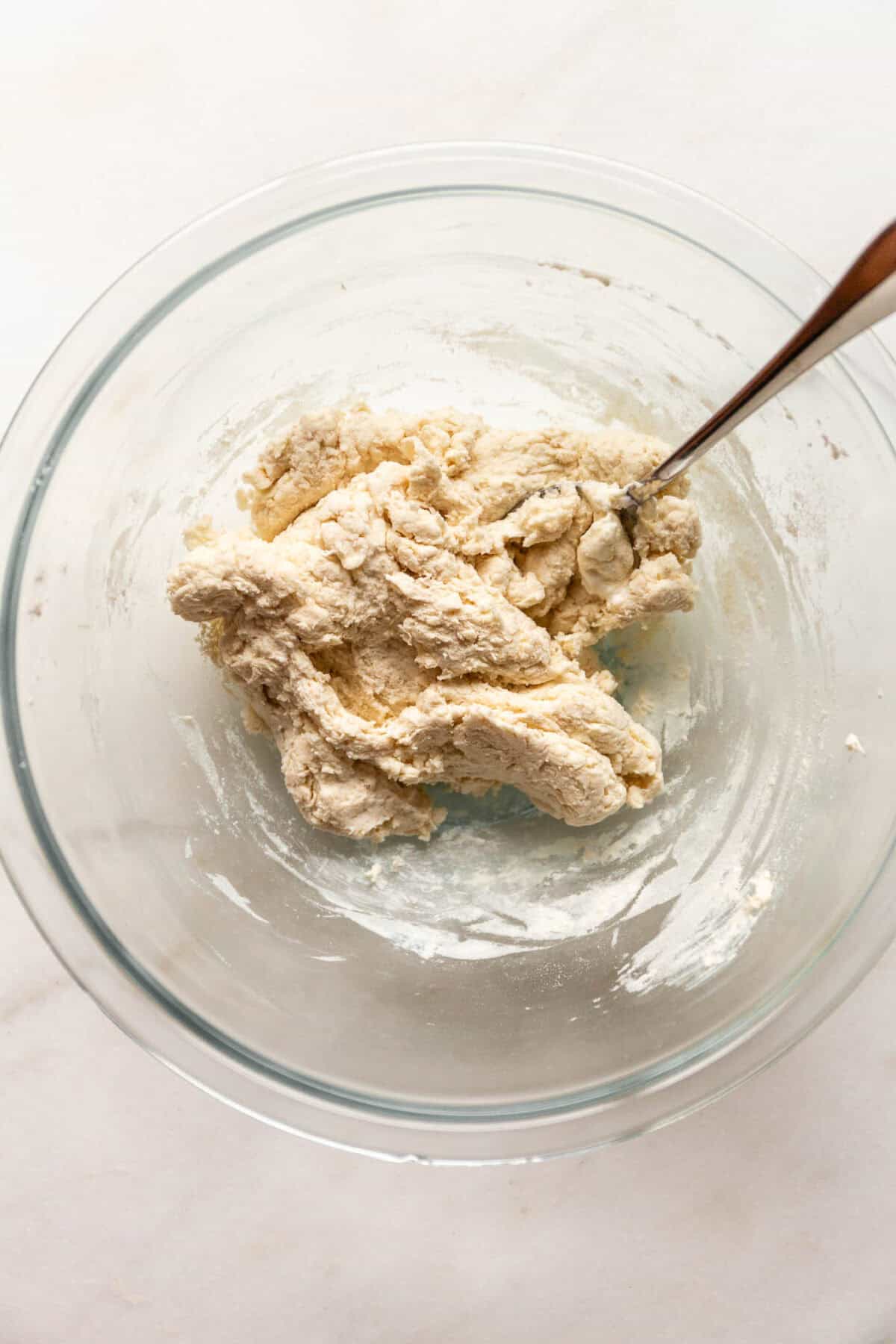 A glass bowl of thick, clumpy dough being mixed with a spoon.