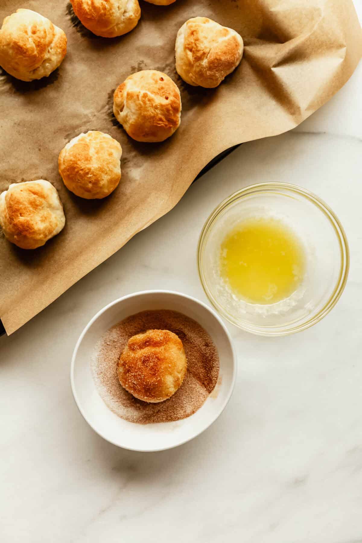 Baked donut holes on parchment paper next to a bowl of melted butter and a bowl of cinnamon sugar with one donut hole being coated.