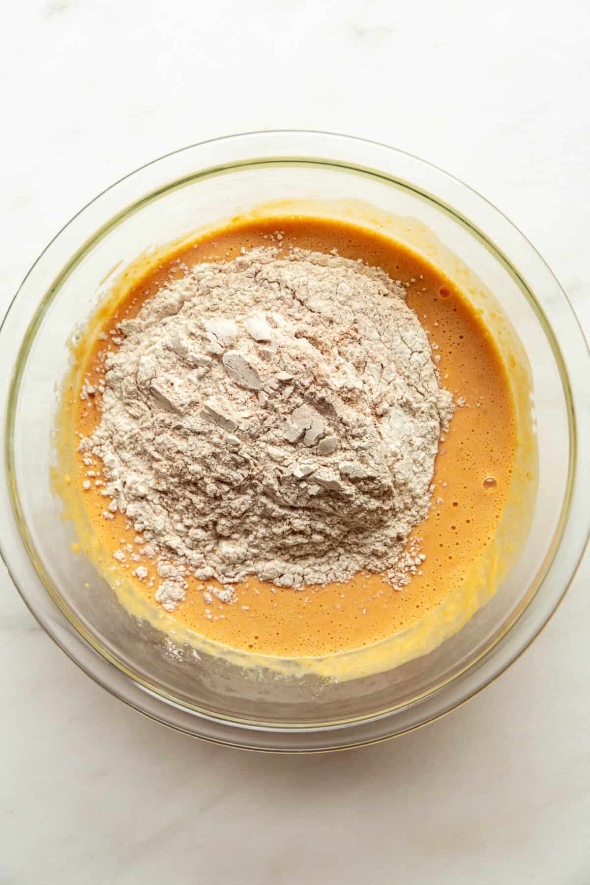 Flour and dry ingredients added on top of pumpkin muffin batter in a glass mixing bowl, not yet stirred in.