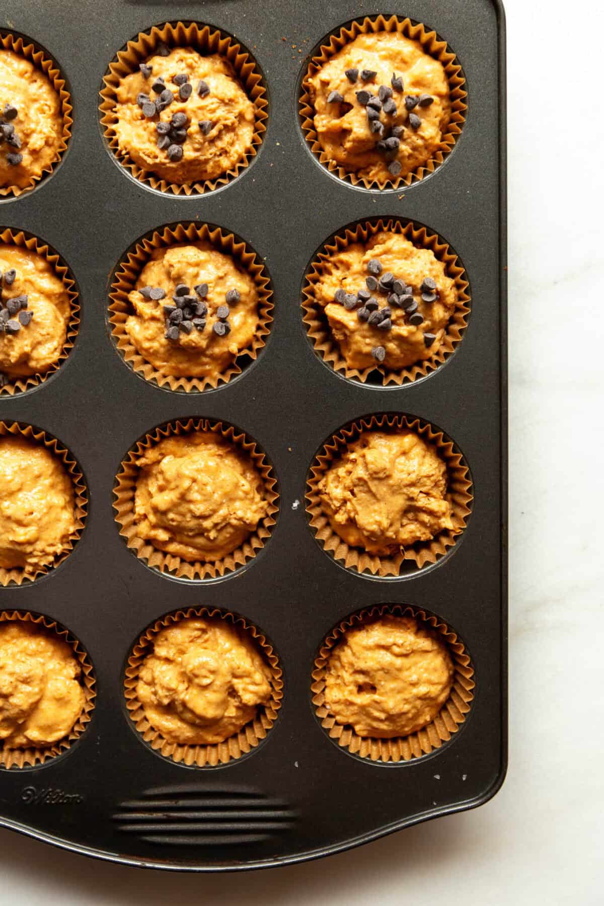 Unbaked cottage cheese pumpkin muffin batter portioned into a muffin tin, some cups topped with chocolate chips.