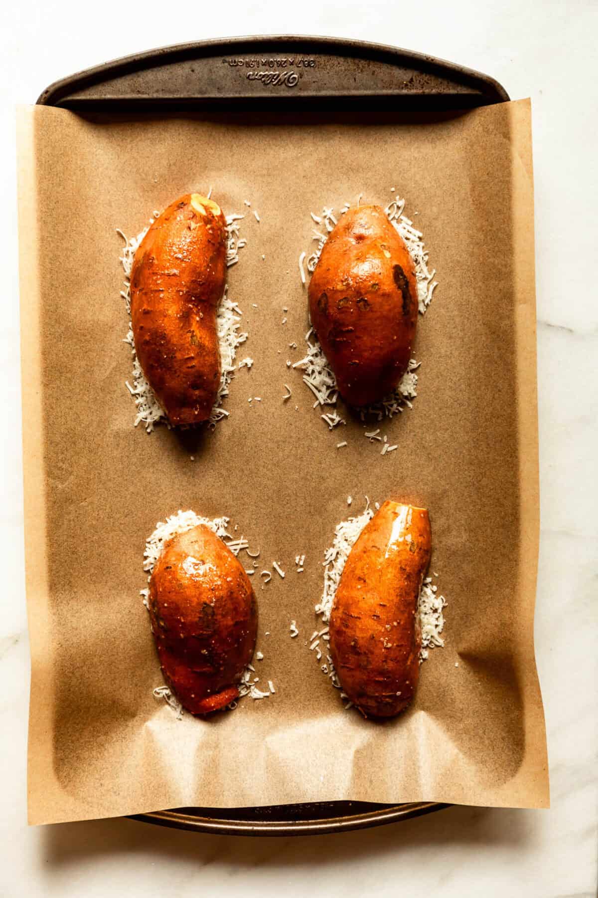 Halved sweet potatoes topped with shredded parmesan cheese on a parchment-lined baking sheet before roasting.