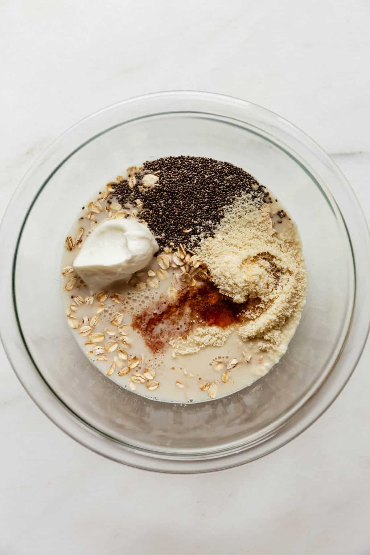 Oats, chia seeds, almond flour, yogurt, and liquid ingredients added to a glass bowl for almond croissant overnight oats.