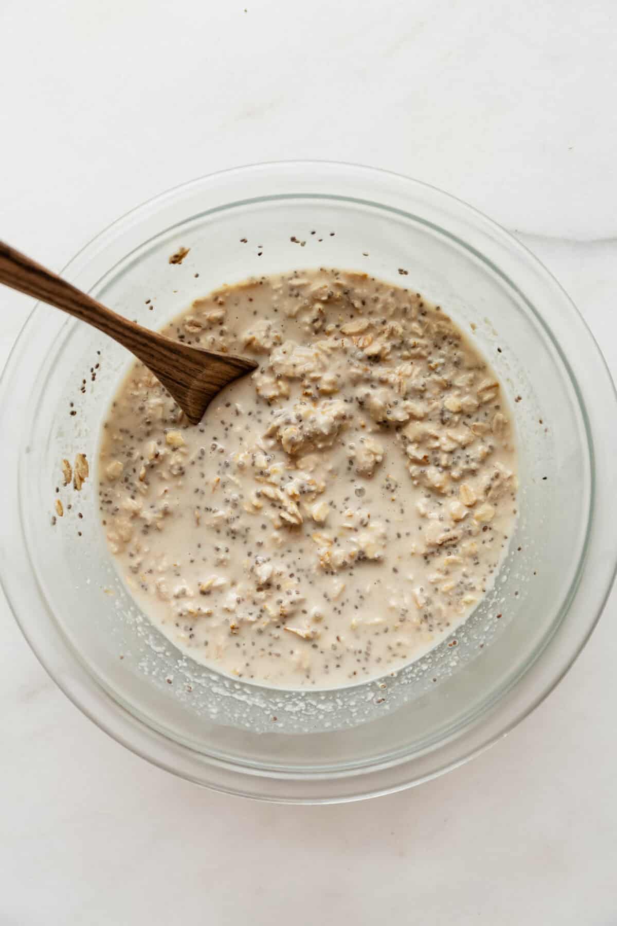 Stirring the almond croissant overnight oats mixture with a wooden spoon in a glass bowl before chilling.