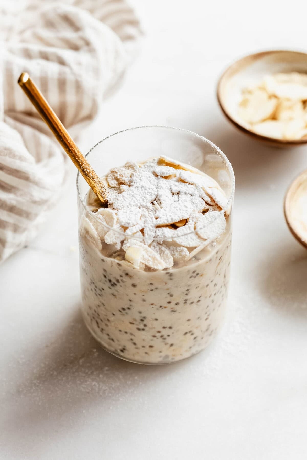 Almond croissant overnight oats in a glass jar topped with sliced almonds and powdered sugar, with a gold spoon and small bowls of almonds on the side.