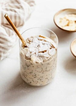 Almond croissant overnight oats in a glass jar topped with sliced almonds and powdered sugar, with a gold spoon and small bowls of almonds on the side.