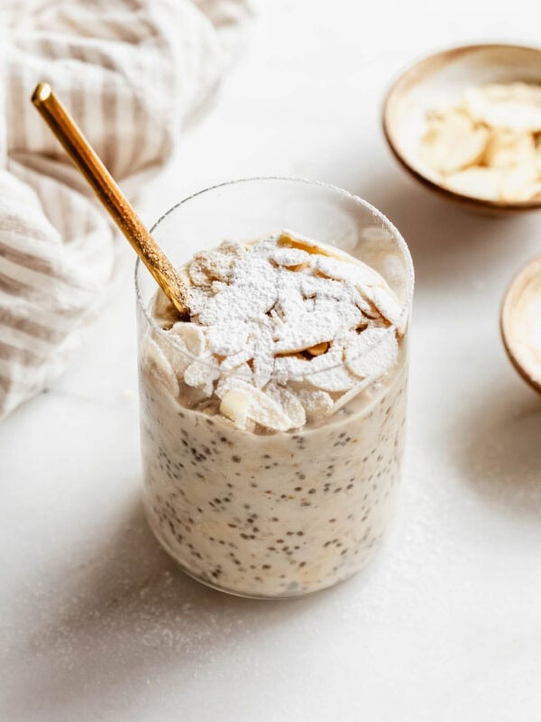 Almond croissant overnight oats in a glass jar topped with sliced almonds and powdered sugar, with a gold spoon and small bowls of almonds on the side.