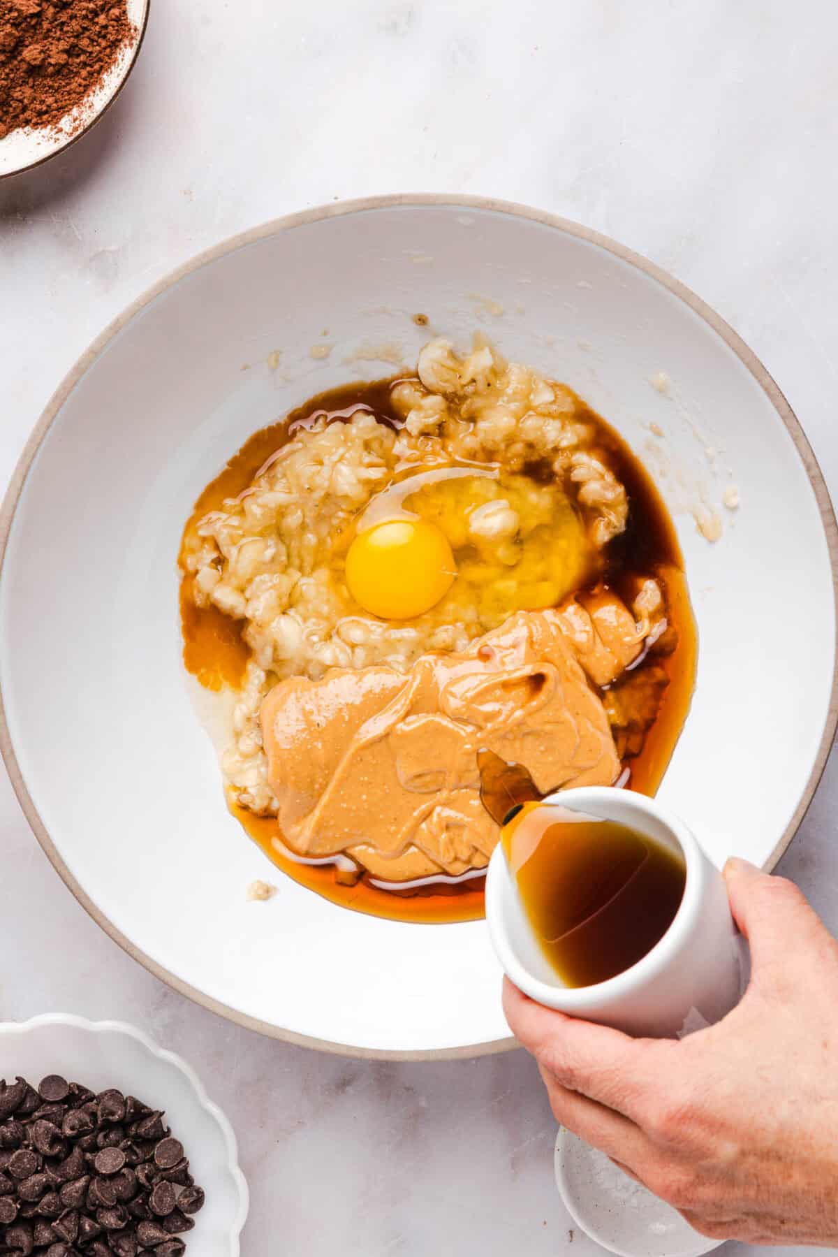 a hand pouring maple syrup into a bowl of mashed banana, peanut butter and egg.