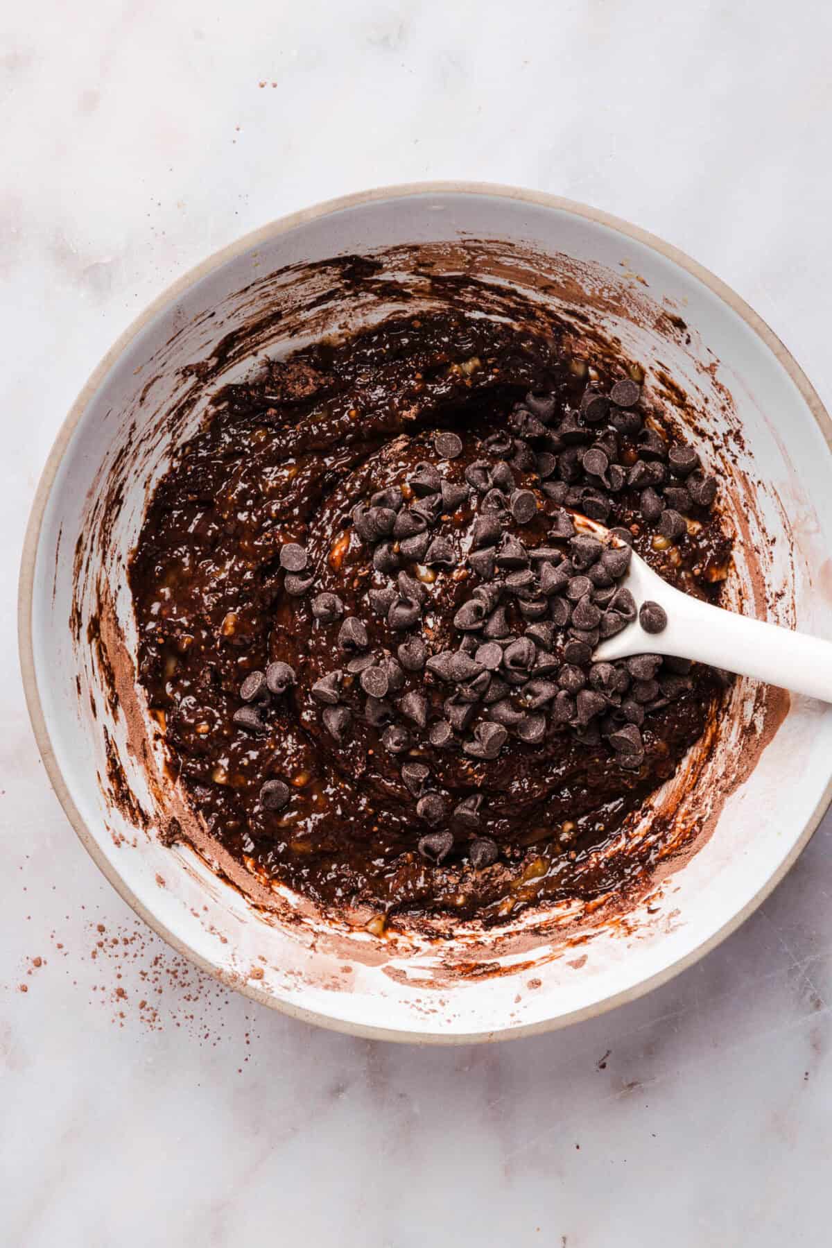 Chocolate chips folded into thick banana brownie batter in a white mixing bowl