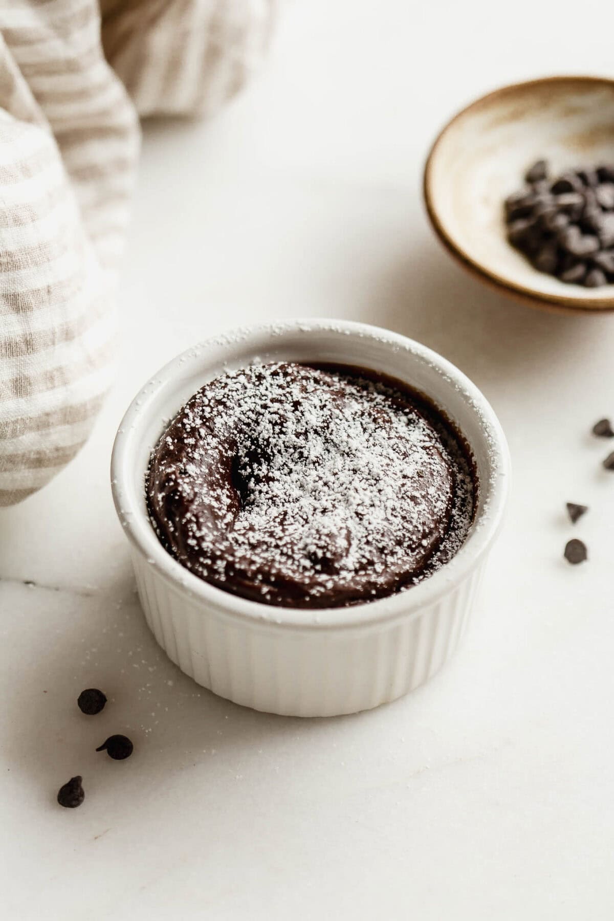 Baked cottage cheese chocolate lava cake dusted with powdered sugar in a white ramekin, with a small bowl of chocolate chips on the side.