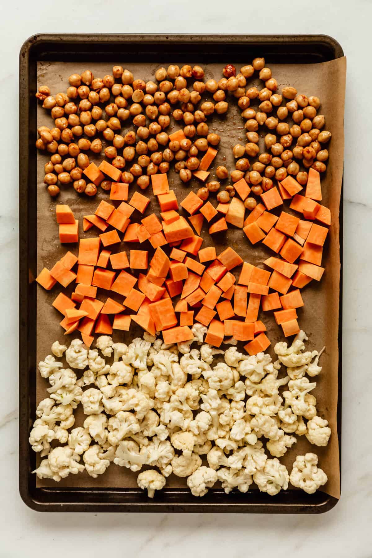 Chickpeas, cubed sweet potato and cauliflower florets arranged in rows on a parchment-lined baking sheet, ready to roast for fall harvest quinoa salad.