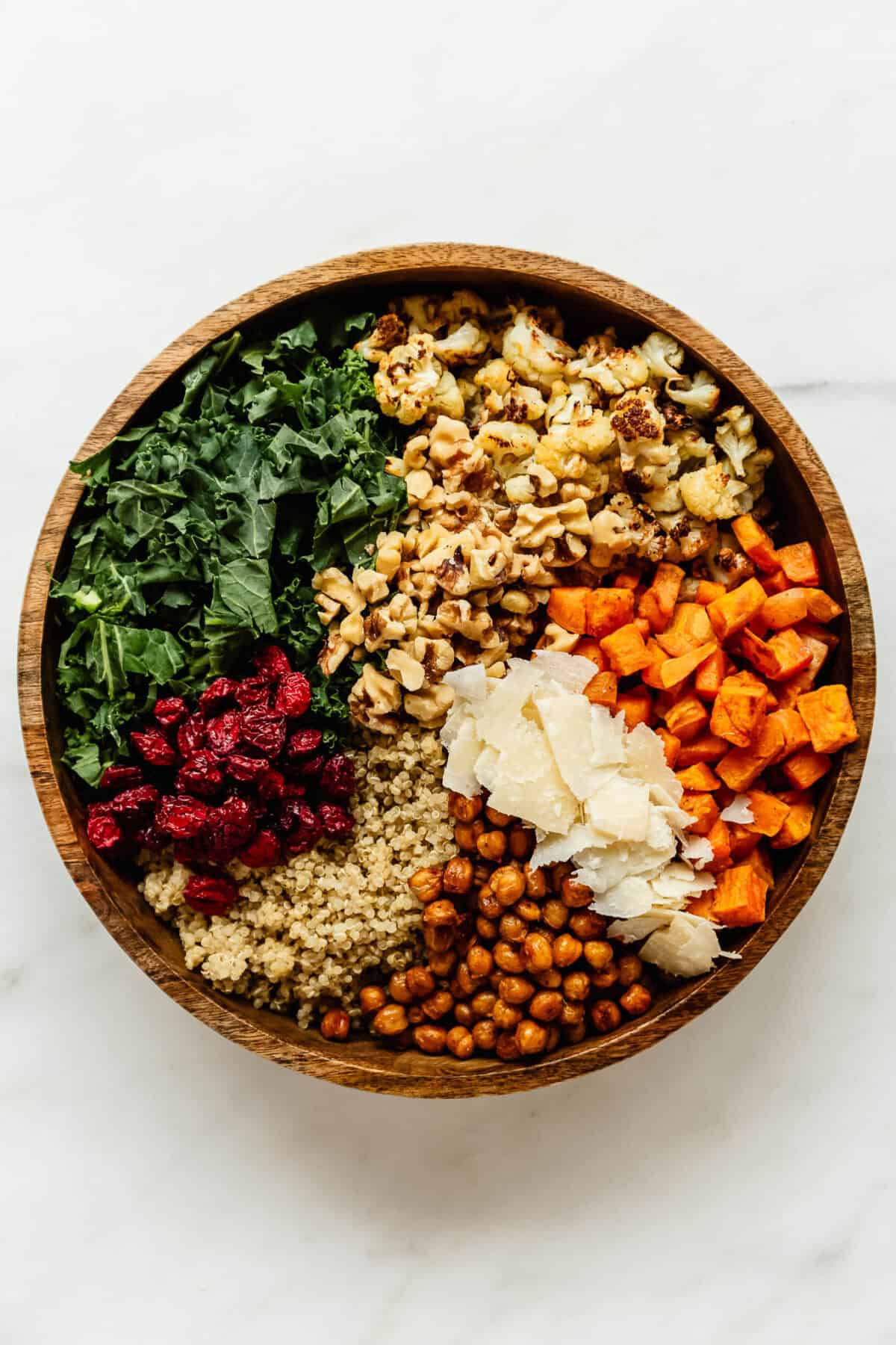 Overhead view of fall harvest quinoa salad ingredients arranged in sections in a wooden bowl, including kale, roasted cauliflower, walnuts, roasted sweet potato, shaved parmesan, crispy chickpeas, quinoa and dried cranberries.