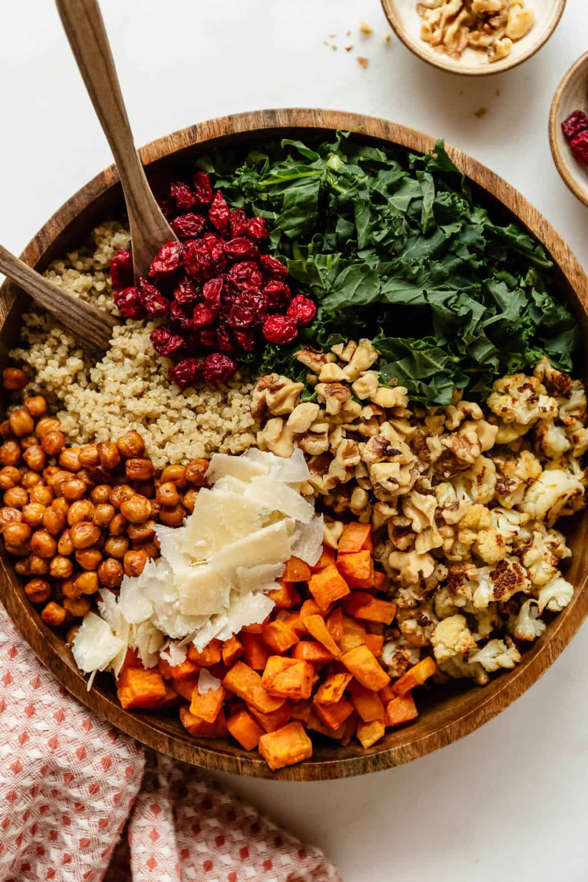 Fall harvest quinoa salad in a wooden bowl with wooden serving spoons, topped with kale, dried cranberries, quinoa, crispy chickpeas, shaved parmesan, roasted sweet potato, roasted cauliflower and walnuts.