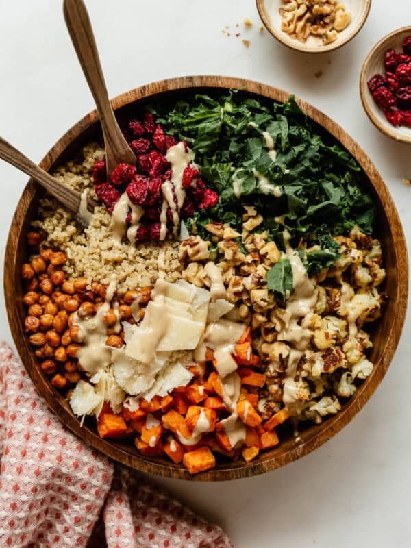 Fall harvest quinoa salad drizzled with creamy dressing in a wooden bowl, with roasted sweet potato, crispy chickpeas, kale, cranberries, walnuts and shaved parmesan, plus small bowls of walnuts and cranberries on the side.