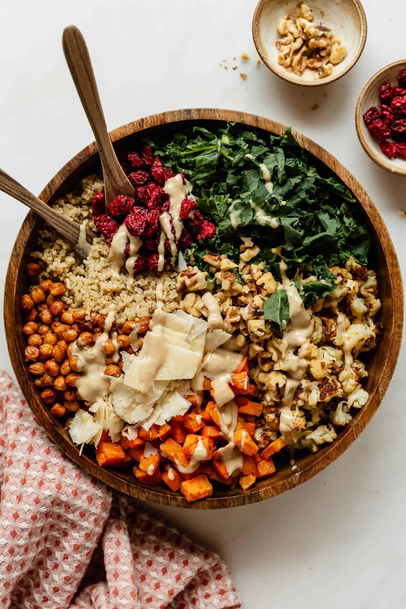 Fall harvest quinoa salad drizzled with creamy dressing in a wooden bowl, with roasted sweet potato, crispy chickpeas, kale, cranberries, walnuts and shaved parmesan, plus small bowls of walnuts and cranberries on the side.