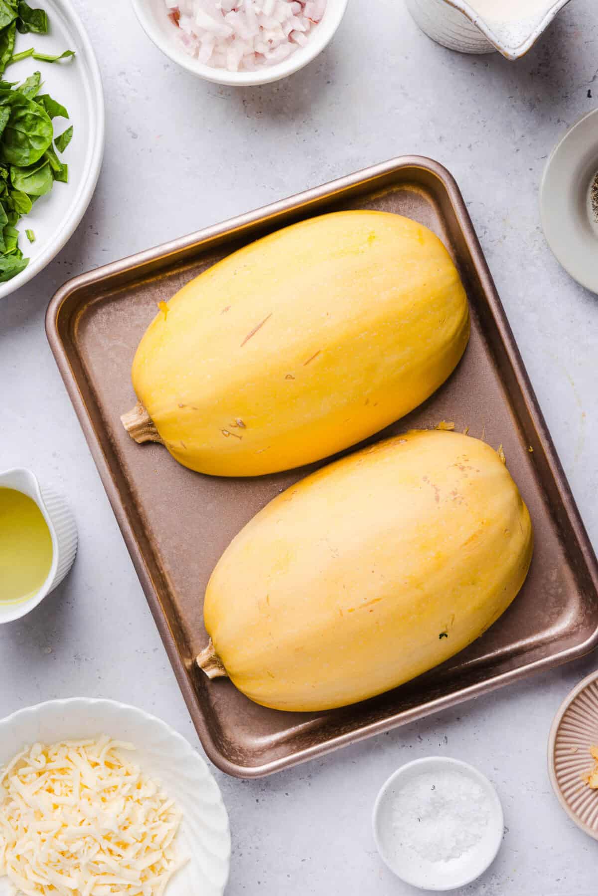 Two roasted spaghetti squash halves cut side down on a sheet pan, surrounded by ingredients